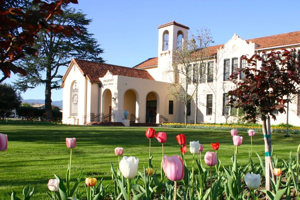 The building is labeled "Fremont High School." The image shows a beautiful school building with Spanish-style architecture, featuring a white facade, a bell tower, and arched doorways. The lawn in front is well-maintained, and colorful tulips in red, white, pink, and yellow are blooming in the foreground, adding vibrancy to the scene. Tall trees, including one with dark red leaves, frame the image, and the sky is clear, creating a bright and inviting atmosphere. 